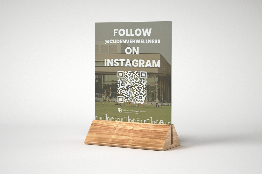A physical mockup of a CU Denver Wellness promotional flyer featuring a QR code, displayed in a wooden desk stand.